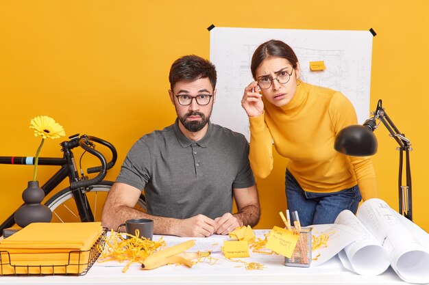 Conceito de trabalho de ocupação de cooperação de pessoas. Confundidos e surpresos, colegas de trabalho, mulher e homem que trabalham duro posam para a mesa com papéis animados com o resultado final, desenhe esboços durante o dia de trabalho no escritório