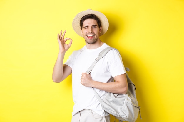 Conceito de turismo, viagens e férias. Feliz turista vai sair de férias, segurando a mochila e mostrando o sinal de tudo bem, sorrindo satisfeito, em pé contra um fundo amarelo.