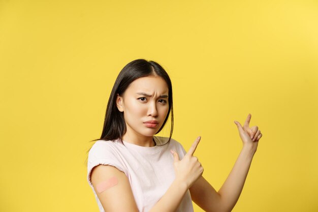 Conceito de vacinação Covid-19. Menina asiática amuada e desapontada apontando para um banner promocional sendo vacinada contra coronavírus, fundo amarelo