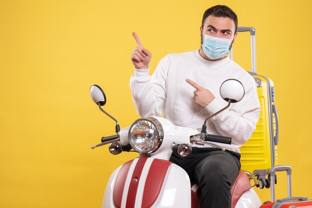 Conceito de viagem com um cara curioso com máscara médica sentado em uma motocicleta com uma mala amarela nela