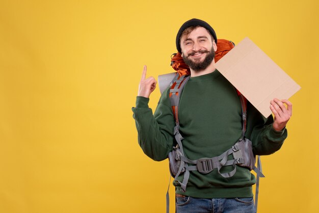 Conceito de viagens com um jovem sorridente com packpack e apontando o espaço livre para escrever apontando para cima no amarelo