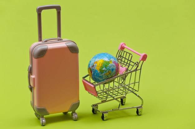 Conceito de viagens e compras. mini mala de viagem de plástico e carrinho de supermercado com globo sobre fundo verde.