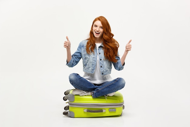 Conceito de viagens e estilo de vida. Jovem mulher caucasiana animada sentado na mala de bagagem aparecendo o polegar. Isolado no branco. Pronto para férias.