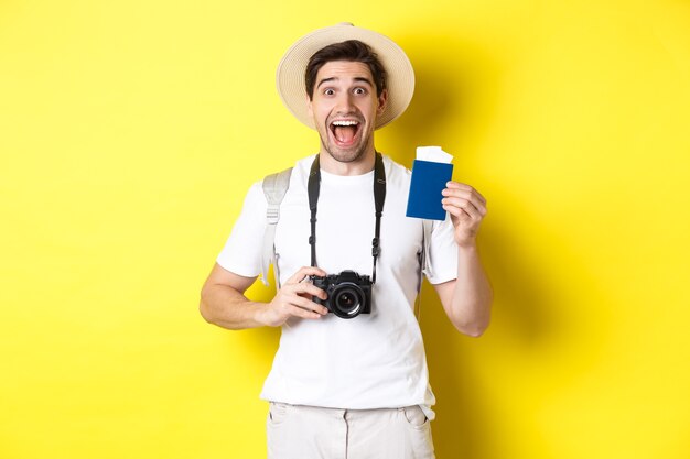 Conceito de viagens, férias e turismo. Tpirost empolgado mostrando passaporte com ingressos, segurando a câmera e usando chapéu de palha, em pé sobre um fundo amarelo.