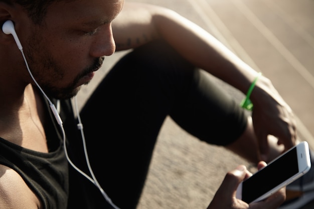 Foto grátis conceito humano e tecnologia. pessoas e esporte. bonitão africano em fones de ouvido, ouvindo música usando seu telefone inteligente com tela de espaço em branco da cópia para o seu texto de publicidade ou informações