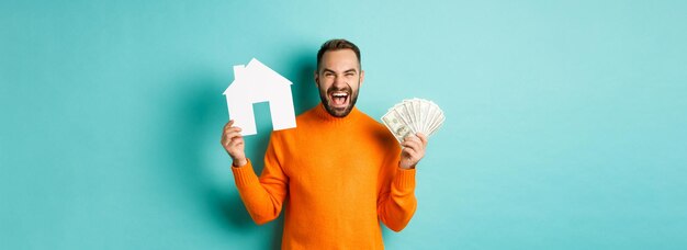Conceito imobiliário e conceito de hipoteca homem caucasiano em êxtase segurando dinheiro dólares e casa faz