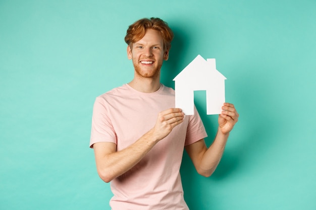 Conceito imobiliário. Jovem com cabelo ruivo, vestindo camiseta, mostrando o recorte da casa de papel e sorrindo feliz, em pé sobre o fundo da casa da moeda.