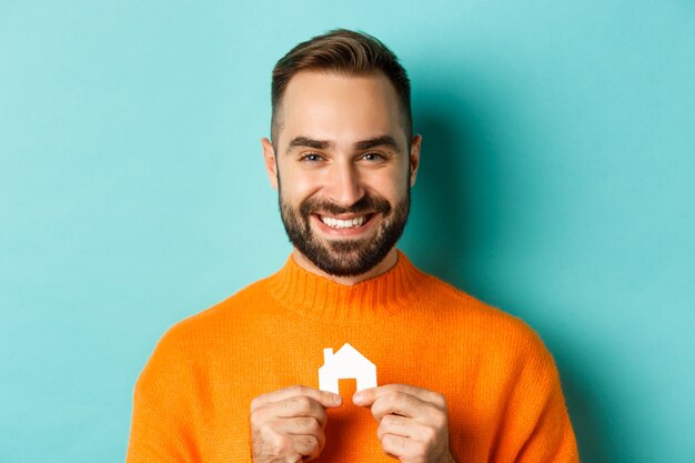 Conceito imobiliário. Jovem feliz procurando aluguel de casa, segurando uma casa de papel sorrindo