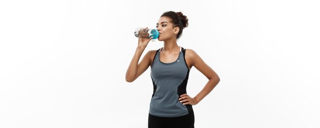 Conceito saudável e fitness linda garota afro-americana em roupas esportivas bebendo água após o treino Isolado no fundo branco do estúdio