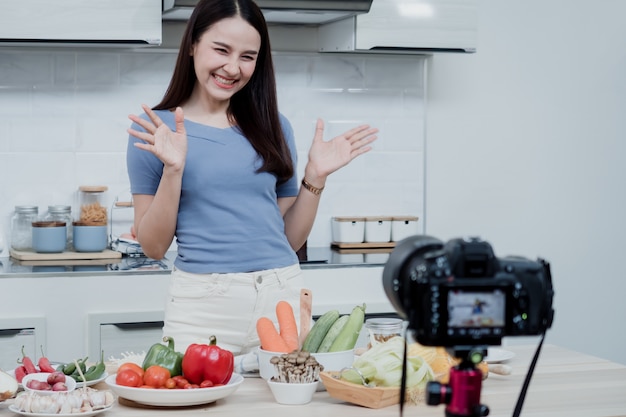 Conceitos de mídia social uma mulher feliz em pé na cozinha usando uma câmera e gravando vídeo online vlogger de mulher asiática feliz transmitindo vídeo online ao vivo ensinando a cozinhar na cozinha em casa.