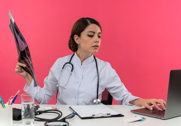 Foto grátis concentrada jovem médica vestindo bata médica e estetoscópio sentada na mesa com ferramentas médicas e laptop segurando uma foto de raio-x usando laptop