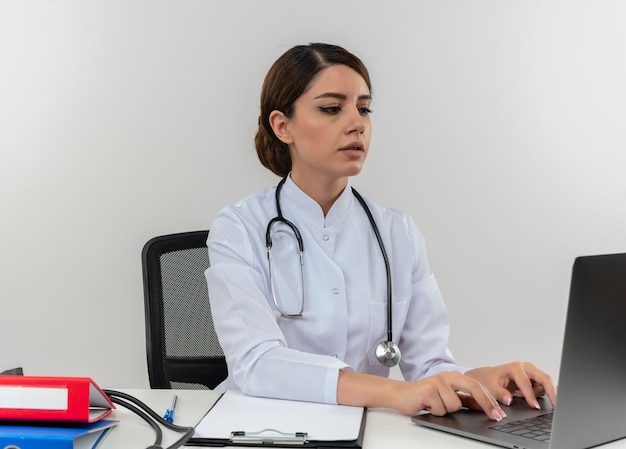 Concentrada jovem médica vestindo túnica médica e estetoscópio, sentada à mesa com ferramentas médicas, usando laptop isolado na parede branca