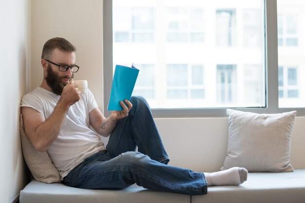 Concentrado homem de óculos, bebendo café enquanto lê o livro