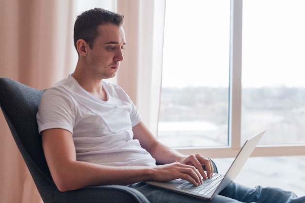 Concentrado homem sentado no chaor e digitando no laptop perto da janela