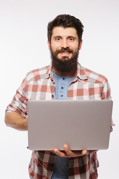 Concentrado jovem barbudo de óculos, vestido com camisa, usando laptop isolado sobre uma parede branca.