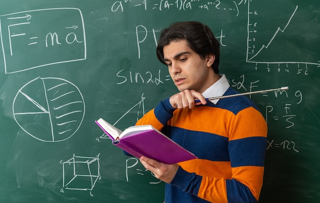 Concentrado jovem professor de geometria em pé em vista de perfil na frente do quadro-negro em sala de aula lendo livro apontando para o lado