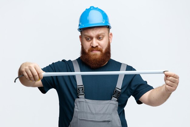 Concentrado jovem trabalhador da construção civil usando capacete de segurança e uniforme segurando e olhando para fita métrica isolada no fundo branco