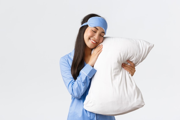 Foto grátis concernente sonhadora linda menina asiática de pijama azul e máscara de dormir deitada na cama com os olhos fechados e abraçando o travesseiro, sorrindo despreocupada como tendo uma boa noite de sono, fundo branco de pé.