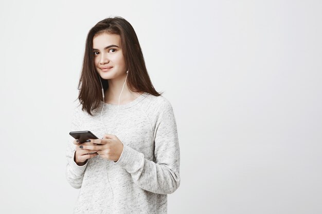 Concurso feminino atraente, com belos olhos e sorriso misterioso, segurando o telefone enquanto escuta música em fones de ouvido