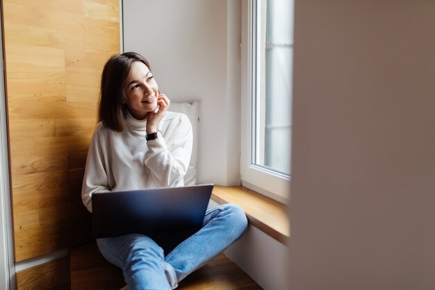 Concurso mulher está trabalhando em seu laptop enquanto está sentado na grande janela em tempo diário