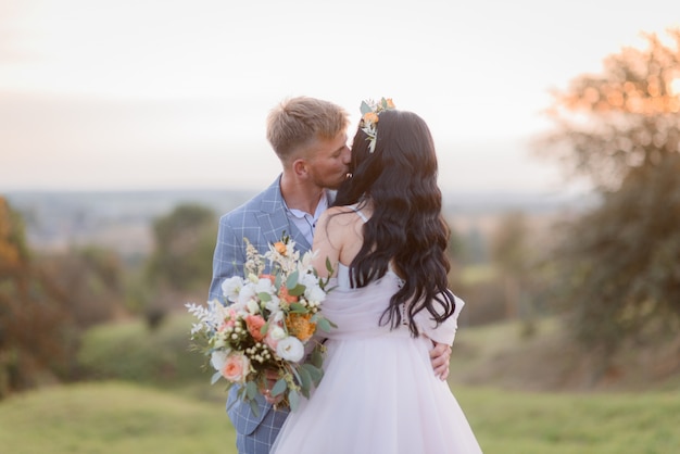 Concurso noiva e noivo estão beijando ao ar livre à noite no prado com buquê de casamento bonito