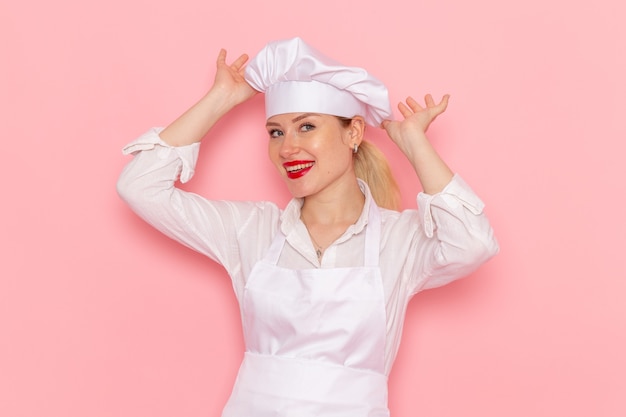 Foto grátis confeitaria feminina de vista frontal, vestida de branco, sorrindo e posando na mesa rosa claro confeitaria trabalho de pastelaria doce