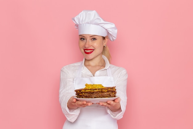 Foto grátis confeiteira feminina em roupa branca segurando deliciosos pastéis sorrindo na parede rosa confeitaria de frente