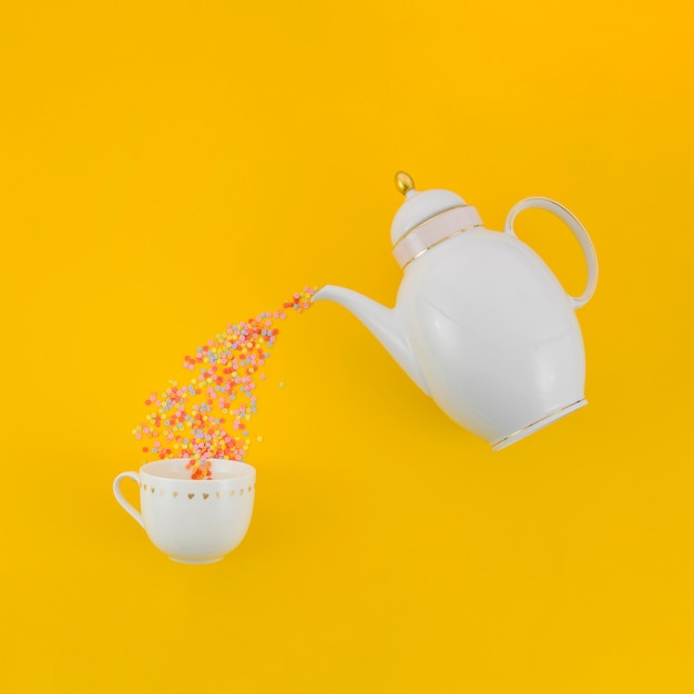 Confetes coloridos derramando de bule de chá branco no copo cerâmico contra fundo amarelo