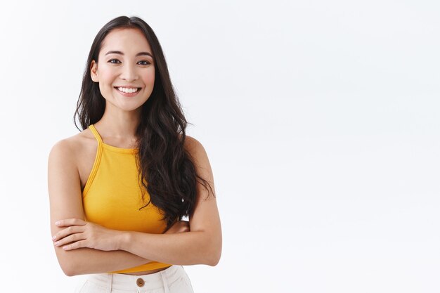 Confiante, atraente e extrovertida mulher asiática em um top amarelo, sorrindo, amigável e feliz enquanto cruza as mãos no peito, posando sobre um fundo branco autoconfiante, pose atrevida, parece determinado