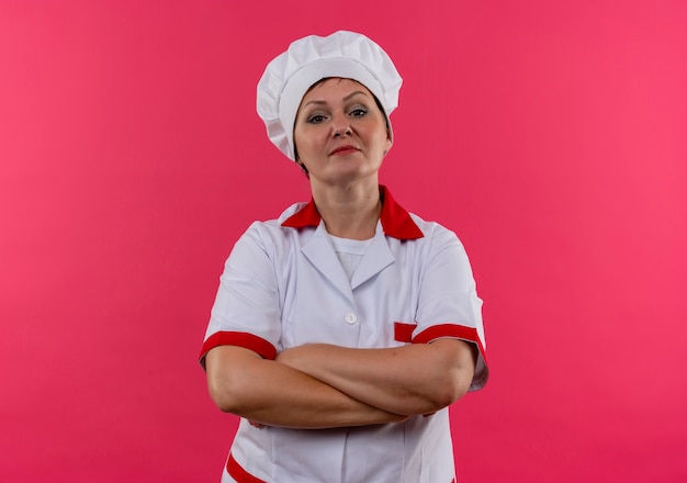 Confiante cozinheira de meia-idade com uniforme de chef, cruzando as mãos na parede rosa isolada com espaço de cópia