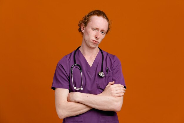 Confiante cruzando as mãos jovem médico masculino vestindo uniforme com estetoscópio isolado em fundo laranja