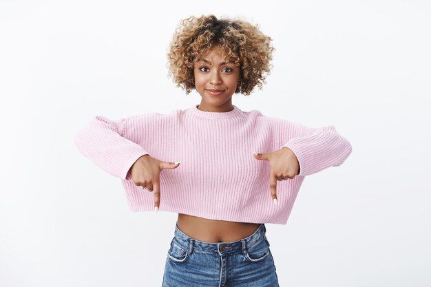 Foto grátis confiante e carismática atraente afro-americana esbelta com corte de cabelo louro apontando para baixo com dedos levantados, sorrindo autoconfiante e dando direção assertiva, mostrando incrível promoção