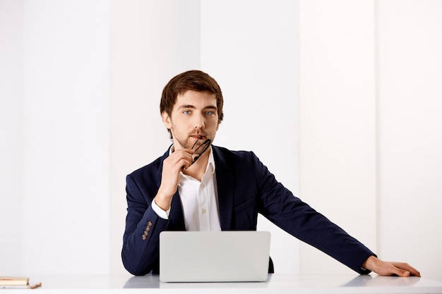 Confiante, elegante empresário de terno, sente-se a mesa de escritório perto de laptop, pensativo