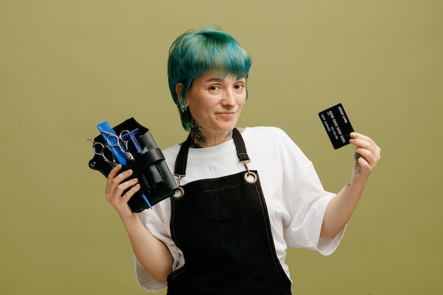 Confiante jovem barbeiro feminino vestindo uniforme olhando para câmera mostrando saco de barbeiro com ferramentas de barbeiro e cartão de crédito isolado em fundo verde oliva