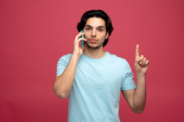 confiante jovem bonito falando no telefone apontando para cima isolado em fundo vermelho com espaço de cópia