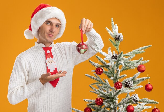 confiante jovem bonito usando chapéu de Natal e gravata de Papai Noel em pé perto da árvore de Natal decorada, segurando e apontando com a mão para o enfeite de bola de Natal, parecendo isolado na parede laranja