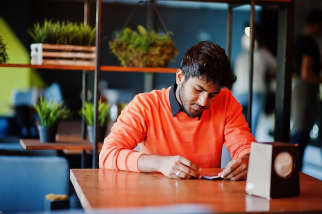 Confiante jovem indiano em roupas casuais inteligentes como suéter laranja sentado no café e usando seu telefone celular
