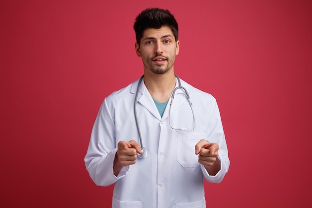 Confiante jovem médico masculino vestindo uniforme médico e estetoscópio no pescoço, olhando para a câmera, mostrando-lhe o gesto isolado no fundo vermelho