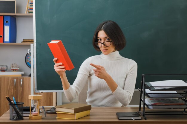 confiante jovem professora usando óculos segurando e aponta para o livro sentado na mesa com ferramentas escolares em sala de aula