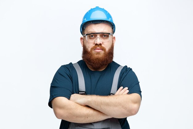 Confiante jovem trabalhador da construção civil vestindo uniforme de capacete de segurança e óculos de segurança olhando para a câmera com os braços cruzados isolados no fundo branco