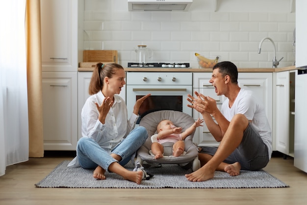 Foto grátis conflito familiar, menino ou menina bonito na cadeira de balanço, pais xingando sentados no chão na cozinha, discutindo perto da filha ou filho recém-nascido, tendo problemas em seu relacionamento.