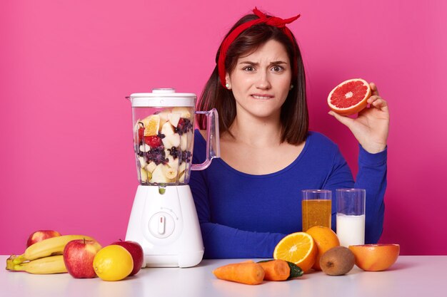 Confusa de cabelo preto emocional fêmea senta-se à mesa, mordendo o lábio, segurando metade de toranja em uma mão, misturando frutas no liquidificador, fazendo doce batido nutritivo para uma refeição saudável na dieta.