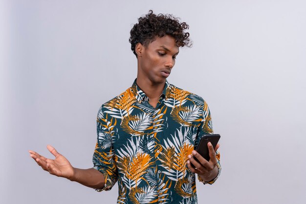 Confuso e surpreso, jovem bonito de pele escura com cabelo encaracolado segurando um telefone celular, tendo dúvidas enquanto levanta a mão