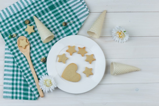 Foto grátis conjunto de cones de waffle, flores e biscoitos no prato e colher de pau no fundo de madeira e toalha de cozinha. colocação plana.