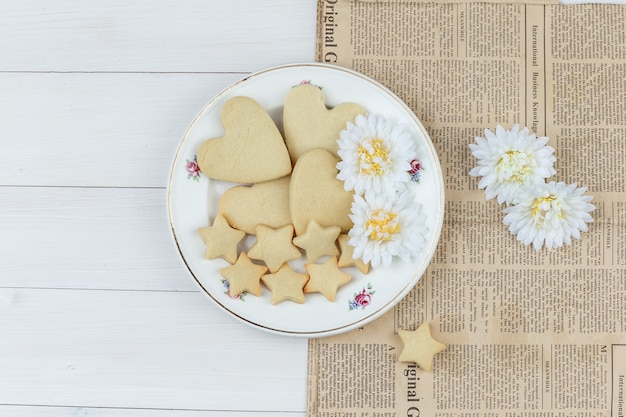 Conjunto de flores e biscoitos em um prato de fundo de madeira e jornal. colocação plana.