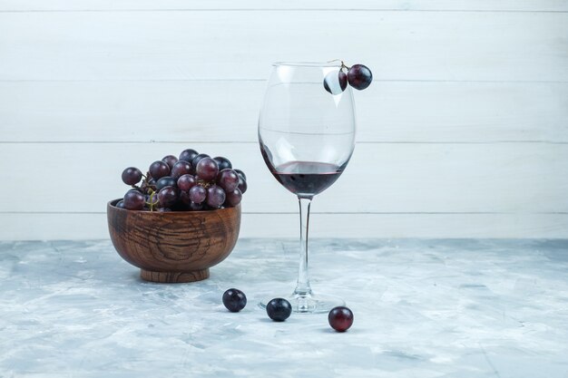 Conjunto de um copo de vinho e uvas pretas em uma tigela de barro no fundo cinza e de madeira sujo. vista lateral.
