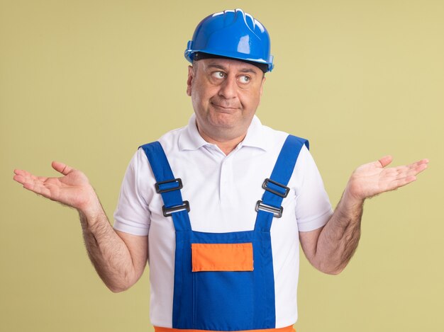 Construtor adulto confuso, de uniforme com as mãos abertas olhando para o lado isolado na parede verde oliva