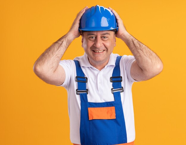 Construtor adulto e sorridente, de uniforme, coloca as mãos na cabeça isolada na parede laranja