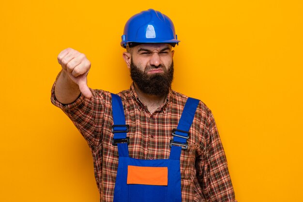 Construtor barbudo com uniforme de construção e capacete de segurança, olhando para a câmera descontente, mostrando os polegares para baixo em pé sobre um fundo laranja