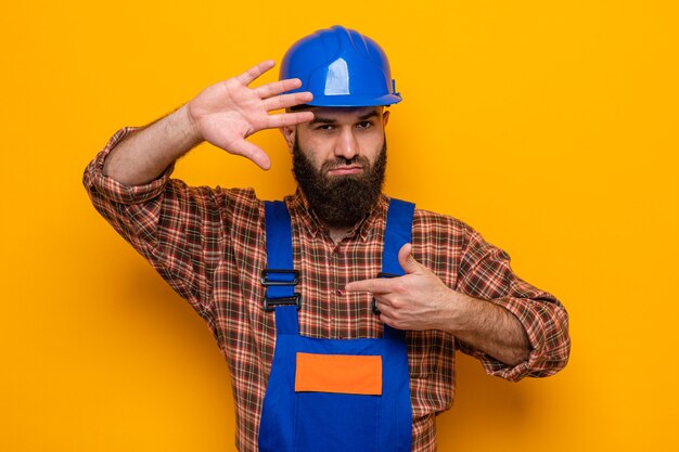 Construtor barbudo com uniforme de construção e capacete de segurança olhando para a câmera, fazendo moldura com as mãos olhando para a câmera através desta moldura em pé sobre um fundo laranja